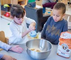 Photo of children using mixing bowls and measuring cups to make a mixture of something.