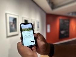 Two hands cradle an iPhone with the University Galleries' digital guide loaded on the screen. In the background, on the right, are framed photos on a white wall, and on the left, a black and red wall with fake weapons.